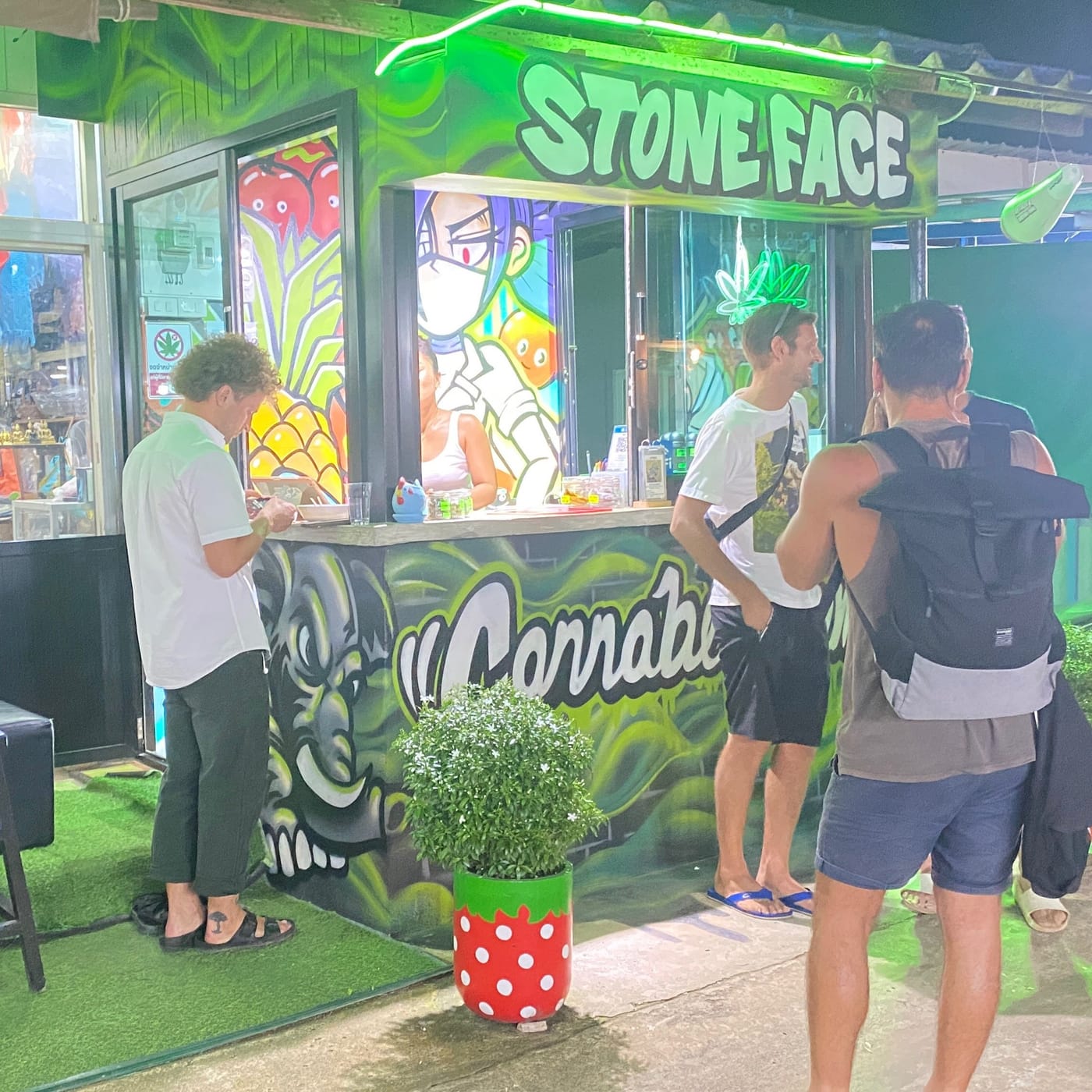 Customers at the counter under the Stone Face sign, beside a graffiti mural of a stone face and cannabis leaves