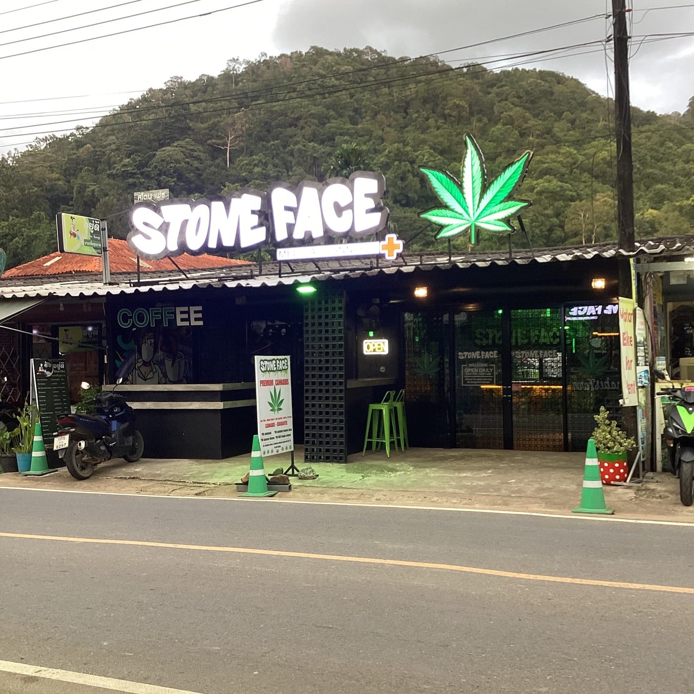 Stone Face from the road in daylight — the lit sign and neon cannabis leaf above an open-fronted shop, COFFEE painted on the left wall, forested hills behind